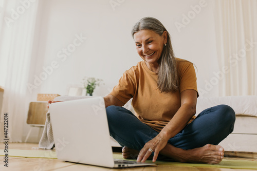 Indoor image of beautiful energetic female on retirement sitting barefoot on floor using laptop turning on yoga video tutorial. Elderly European woman surfing internet on portable computer