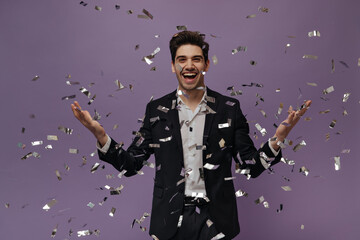 Gorgeous young brunette-haired man in white shirt and black suit smiling and posing with confetti against violet background 