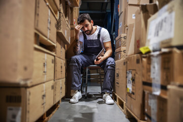 Wall Mural - Young worried hardworking tattooed bearded blue collar worker in overalls sitting on the chair in storage of import and export firm, holding tablet and touching his head. Is delivery on time?