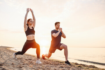 Full body sunlit couple young two friend sporty sportswoman sportsman woman man in sport clothes warm up training do lunges do exercise on sand sea ocean beach outside on seaside in summer day morning