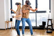 © Roman - Young beautiful caucasian couple playing video games in virtual reality glasses. Funny Man and woman wearing virtual reality goggles for mazing experience in abstract vr world, at home in living room
