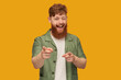 Indoor shot of young handsome redhead man with big beard, wears green t-shirt, looks into camera, wink and points with both hands at you. Isolated over yellow background