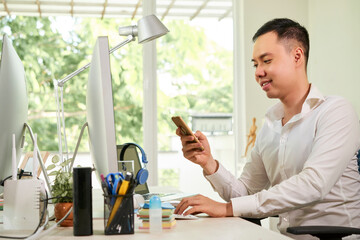 Wall Mural - handsome smiling young businessman sitting at office table and answering text messages on his smartp