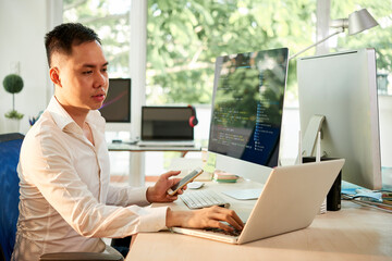 Wall Mural - young software engineer with smartphone in hand working on laptop when checking programming code