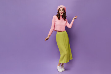 Wall Mural - Pretty happy curly woman dances on purple background. Charming young lady in green midi skirt and pink beret moves and smiles on isolated.