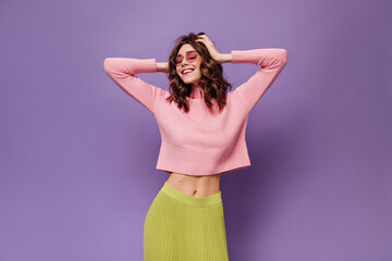 Wall Mural - Curly girl ruffles hair on isolated. Cool charming woman in pink sweater, green skirt and sunglasses smiles on purple background.