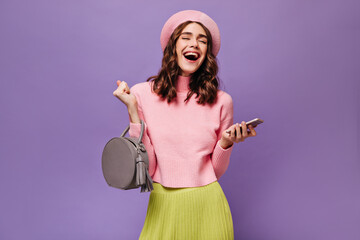 Wall Mural - Joyful woman holds grey handbag and smartphone. Attractive curly girl in pink beret, sweater and green skirt laughs on purple background.
