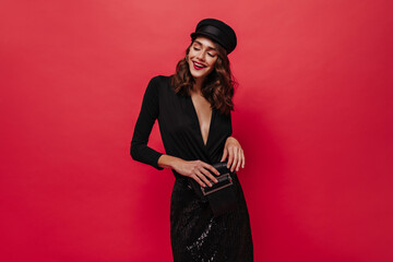 Wall Mural - Excited curly woman in black shiny skirt and cap smiles, holds clutch bag and poses on isolated red background.