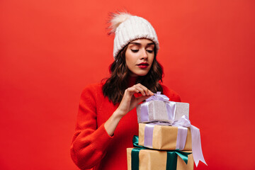 Curly woman with bright lips untie bow on gift box. Cheerful lady in knitted white hat poses on isolated red background.