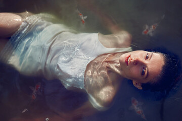 Beautiful young woman in white dress lying in the water with fishes. High angle view