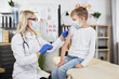 © sofiko14 - Medicine and vaccination concept during pandemic. Beautiful qualified blond woman doctor ready to prick needle into her patient boy's arm. Kid vaccination in medical center