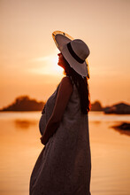 The Silhouette Of A Pregnant Woman At Sunset. The Silhouette Of A Young Woman In A Straw Hat Holding Her Pregnant Belly With Her Hands On The Sea With The Setting Orange Vivid Sun. 