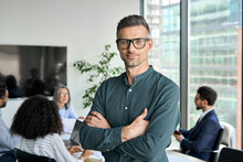 Smiling Confident Mature Businessman Leader Looking At Camera Standing In Office At Team Meeting. Male Corporate Leader Ceo Executive Manager Wearing Glasses Posing For Business Portrait Arms Folded.