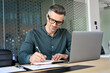 © insta_photos - Happy mature executive ceo manager using laptop writing notes in notebook at workplace. Smiling middle aged business man working in office analyzing financial data doing research.