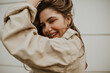 Leinwanddruck Bild - Brown-eyed woman in beige trench coat ruffles hair outside. Happy girl in jacket smiles and poses in good mood.
