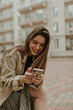 © Look! - Charming brunette woman in checkered grey skirt and beige trench coat sits outside in good mood and chats via phone messenger.