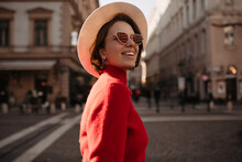Optimistic Beautiful Lady In Beige Hat And Eyeglasses Smiles Outside. Charming Woman In Red Sweater Walks On Street.