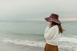 © Look! - Brunette woman looks away at sea. Young attractive girl in wide-brimmed hat, brown pants and sweatshirt poses at beach.