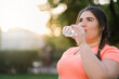 © golubovy - Water freshness. Energy recovery. Fitness well-being. Body positive. Joyful thirsty obese overweight woman drinking clear liquid in defocused copy space sunset landscape.
