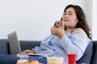 © Bangkok Click Studio - Asian young happy beautiful fat female shopper sit relaxing on fabric sofa look at camera using laptop computer searching products shopping online while eating delicious chocolate donut in hand