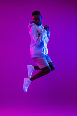 Wall Mural - Full lenght portrait of young energetic African American man jumping in dark purple futuristic cyberpunk neon light background