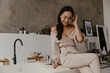 Brunette happy woman dressed in stylish linen pants and beige blouse shyly looks down, smiles and sits on bath in bathroom.
