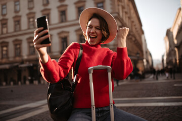 Wall Mural - Joyful short-haired woman in red sweater, beige hat and jeans rejoices, takes selfie, holds smartphone and sits on suitcase outside.