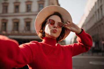 Wall Mural - Short-haired woman in red sweater and sunglasses takes selfie outside. Beautiful lady in beige hat smiles on street.