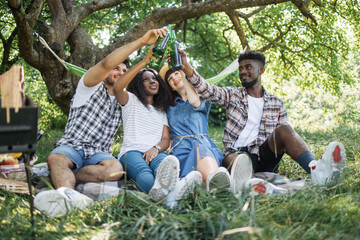 Wall Mural - Multiracial group of friends in summer clothes drinking beer during picnic at green garden. Young happy people relaxing together on nature . Concept of friendship and enjoyment.