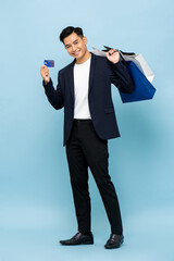 Wall Mural - Full length portrait of handsome southeast Asian millenial man holding shopping bags and credit card in isolated studio light blue background for cashless payment concept