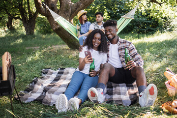 Wall Mural - Lovely black couple sitting in hugs at garden and looking on mobile screen while their diverse friends talking and drinking in hammock. Four friends having picnic on nature.