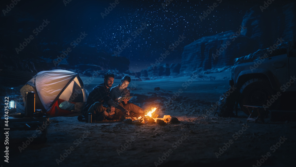 Happy Couple Nature Camping in the Canyon at Night, Use Digital Tablet Computer, Sitting by Campfire. Two Traveling people Have Fun, Smile, Post on Social Media, Watch Funny Videos on Internet - obrazy, fototapety, plakaty 