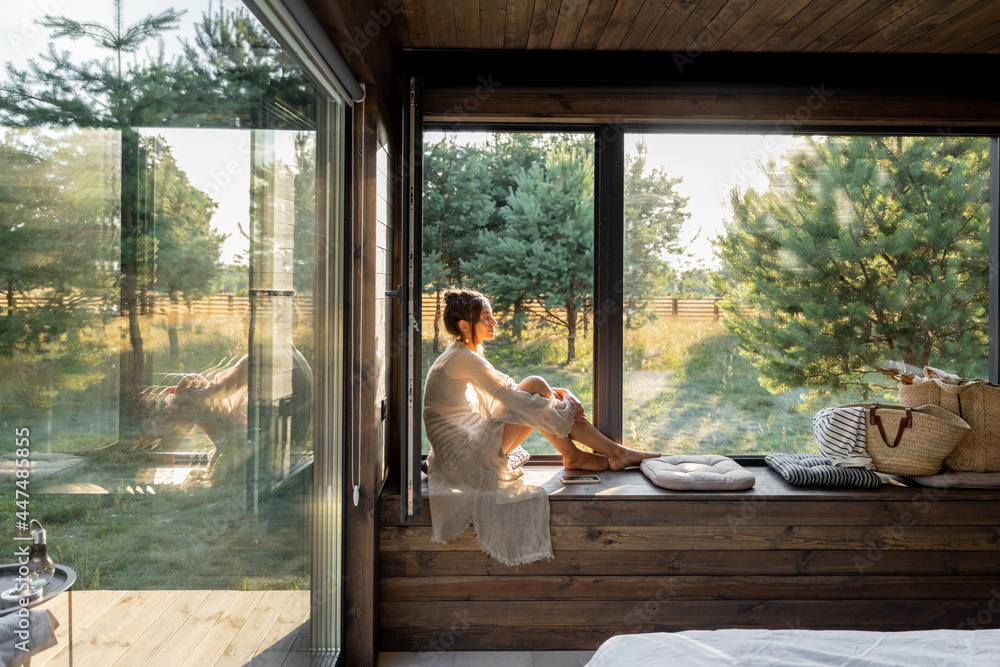 Young woman resting at beautiful country house or hotel, sitting on the window sill enjoying beautiful view on pine forest. Concept of solitude and recreation on nature - obrazy, fototapety, plakaty 