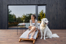 Woman Enjoys The Nature While Sits On Sunbed On Wooden Terrace At The Modern Hotel Resort With Panoramic Windows Near Pine Forest Whith Big White Dog. Concept Of Solitude And Recreation On Nature
