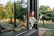 Leinwandbild Motiv Young woman enjoy of resting at modern house or hotel in pine forest, sitting with tablet on the window sill and greeting someone. Beautiful destinations for vacation