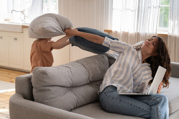 Wall Mural - Irritated mom hitting playful kid with pillow for disturbing from work. Freelancer woman try to use laptop at home sitting on safe in living room with small naughty kid bored. Family on quarantine