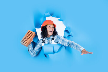 going to throw it. girl builder in hard hat on construction site. engineer hold brick. young female worker on blue background. repair and renovation. woman wear protective helmet