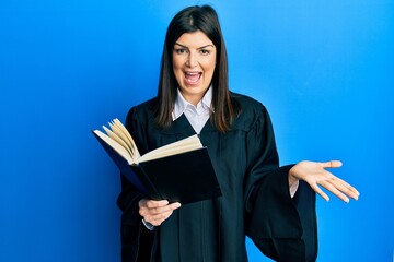 Wall Mural - Young hispanic woman wearing judge uniform reading book celebrating achievement with happy smile and winner expression with raised hand