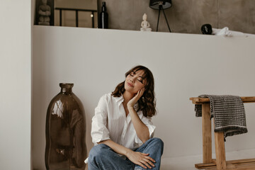 Pretty curly woman in white shirt and jeans sits on floor. Cute pretty girl in cotton blouse and denim pants poses in bathroom.