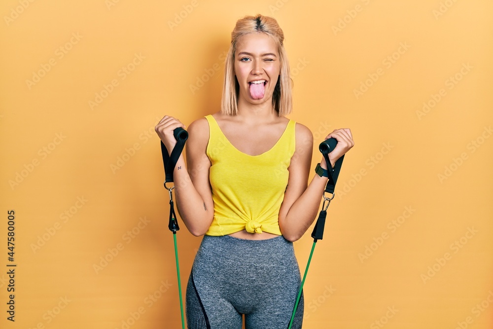Beautiful blonde woman training arm resistance with elastic arm bands sticking tongue out happy with funny expression. - obrazy, fototapety, plakaty 
