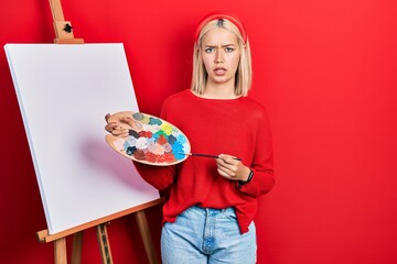 Poster - Beautiful blonde woman standing drawing with palette by painter easel stand clueless and confused expression. doubt concept.