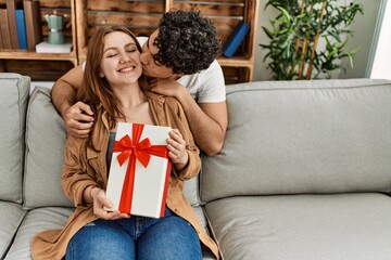 Young couple smiling happy sitting on the sofa surprise with anniversary gift at home.