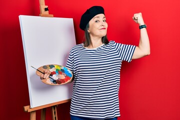 Sticker - Middle age hispanic woman standing drawing with palette by painter easel stand strong person showing arm muscle, confident and proud of power