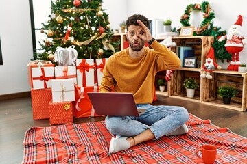 Sticker - Arab young man using laptop sitting by christmas tree doing ok gesture shocked with surprised face, eye looking through fingers. unbelieving expression.