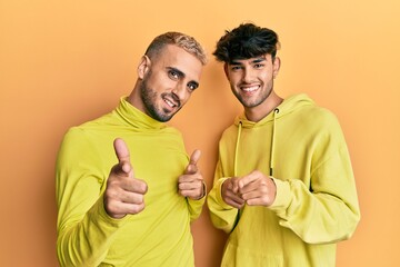 Poster - Homosexual gay couple standing together wearing yellow clothes pointing fingers to camera with happy and funny face. good energy and vibes.