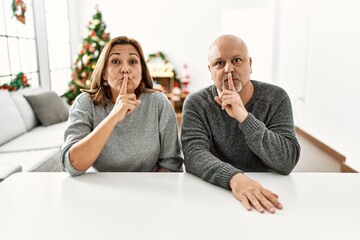 Sticker - Middle age hispanic couple sitting on the table by christmas tree asking to be quiet with finger on lips. silence and secret concept.