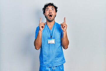 Sticker - Young hispanic man wearing blue male nurse uniform amazed and surprised looking up and pointing with fingers and raised arms.