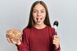 Beautiful brunette little girl eating healthy whole grain cereals holding spoon sticking tongue out happy with funny expression.