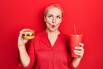 Canvas Print - Young blonde woman eating a tasty classic burger with fries and soda making fish face with mouth and squinting eyes, crazy and comical.