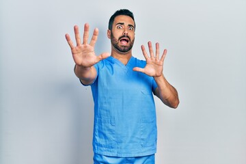 Canvas Print - Handsome hispanic man with beard wearing blue male nurse uniform afraid and terrified with fear expression stop gesture with hands, shouting in shock. panic concept.